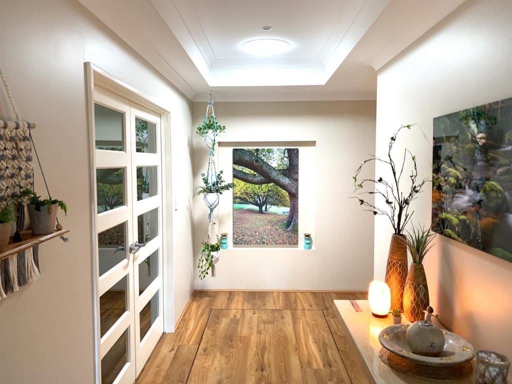 A bright hallway with wooden flooring, white walls, and a large window featuring an outdoor tree view. Shelves hold potted plants, and decorative items are placed on a table with a glowing lamp.