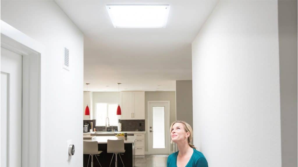 A woman stands in a well-lit hallway looking up at a ceiling light. The hallway leads to an open kitchen area with white cabinetry and red pendant lights.