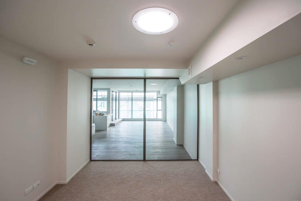 Solatube Skylights: The Secret to a Bright, Beautiful Space 1 A room with beige walls and carpet leads to another room with wooden flooring, separated by large glass doors. A round ceiling light, complemented by a Solatube skylight, illuminates the area.