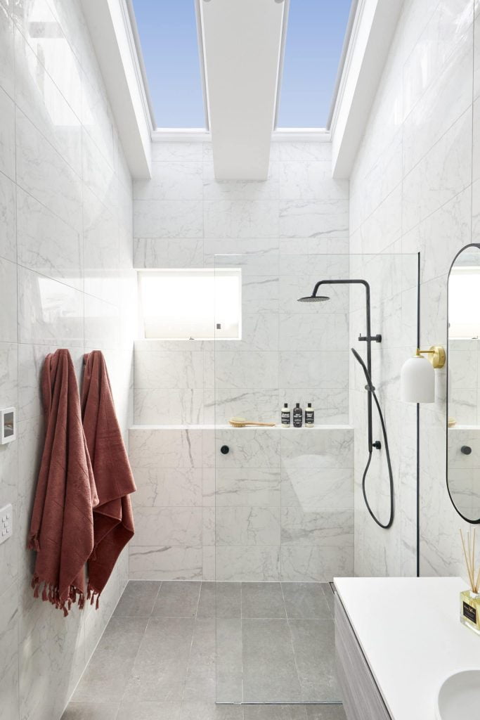 Modern bathroom with white marble tiles, skylights, a glass-enclosed shower featuring black fixtures, wall-mounted soap bottles, two reddish towels hanging on hooks, and a white sink with a mirror. Check the FAQs for more details on this stylish space.