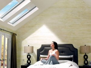A woman sits on a bed in a bright, modern bedroom with skylights that showcase the benefits of skylights for your bedroom, and two bedside lamps.