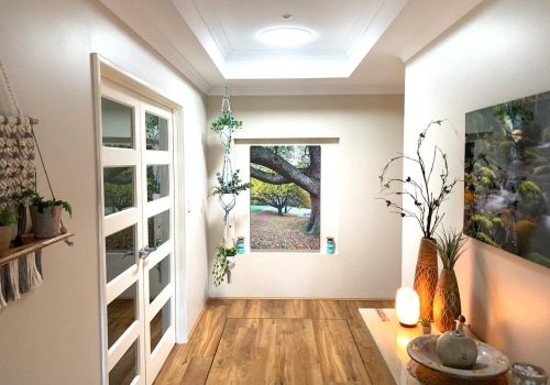 A bright hallway with wooden flooring, white walls, and a large window featuring an outdoor tree view. Shelves hold potted plants, and decorative items are placed on a table with a glowing lamp.