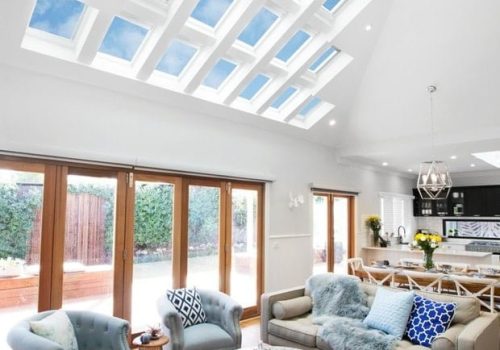 A modern living room with a high ceiling, multiple Velux skylights, and large windows. It includes cozy seating, a coffee table, and an adjacent kitchen area with a dining table. Bright natural light streams in.