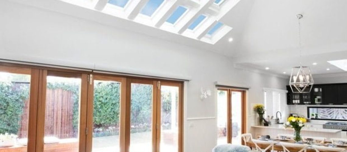 A modern living room with a high ceiling, multiple Velux skylights, and large windows. It includes cozy seating, a coffee table, and an adjacent kitchen area with a dining table. Bright natural light streams in.