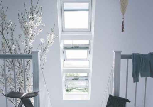 A bright staircase area with multiple skylight windows, a small potted tree with white blossoms, a chair, a hanging decoration, and a folded towel draped over the railing showcases one of the best skylight designs in home renovations.