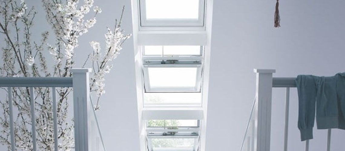 A bright staircase area with multiple skylight windows, a small potted tree with white blossoms, a chair, a hanging decoration, and a folded towel draped over the railing showcases one of the best skylight designs in home renovations.