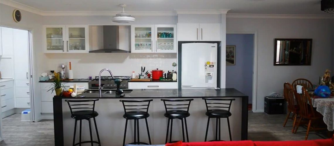 Modern kitchen with a central island featuring bar stools, a white refrigerator, and glass-front cabinets. Velux skylights flood the space with natural light. A dining table with chairs is visible in the adjacent area. Bright and organized space.