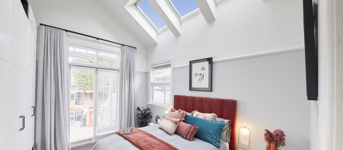 Modern bedroom in Melbourne with a skylight, large windows, and glass doors. The room features a red upholstered bed, bedside tables with lamps, and a mix of gray and red bedding. A framed artwork hangs above the bed.