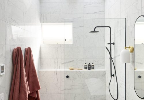 Modern bathroom with white marble tiles, skylights, a glass-enclosed shower featuring black fixtures, wall-mounted soap bottles, two reddish towels hanging on hooks, and a white sink with a mirror. Check the FAQs for more details on this stylish space.