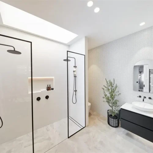 Modern bathroom with a large walk-in shower featuring dual black showerheads, a sleek black vanity with a countertop sink, and a potted plant. Walls are tiled, and lighting is recessed, enhanced by solatubes to brighten the space naturally.