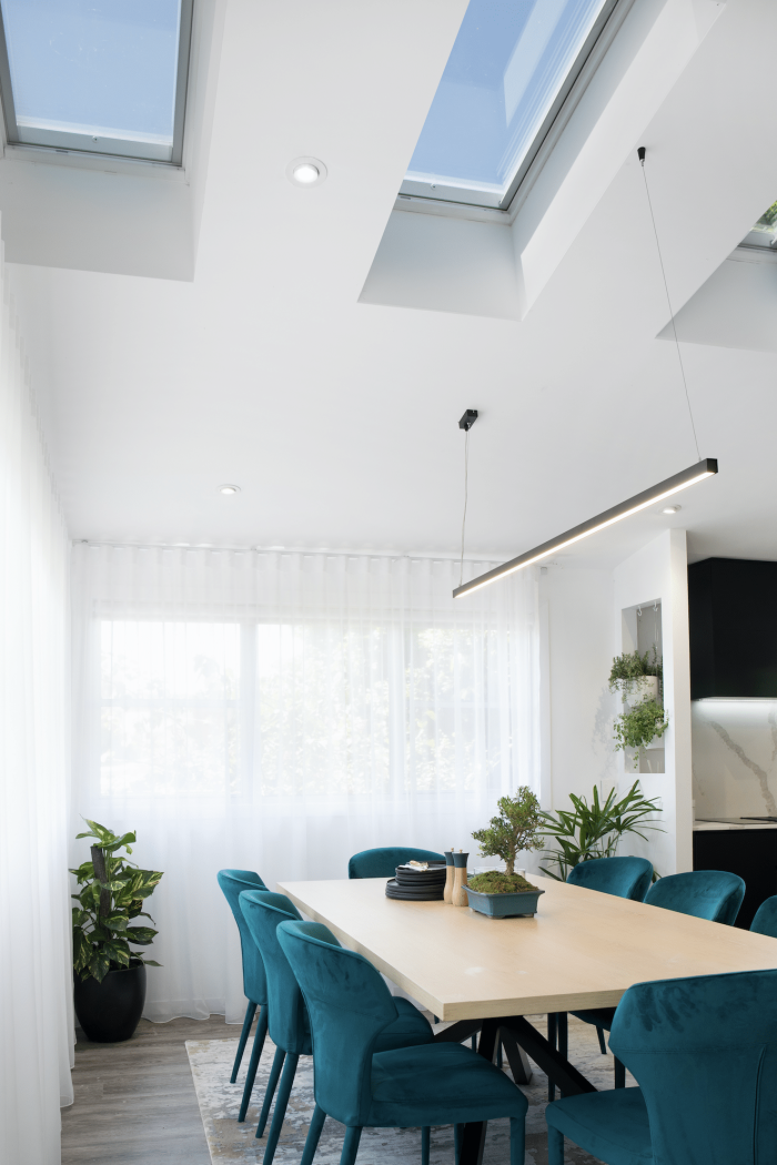 A modern dining area with a light wood table surrounded by teal upholstered chairs. Natural light streams through a Velux skylight and a large window with sheer white curtains. Potted plants are visible.