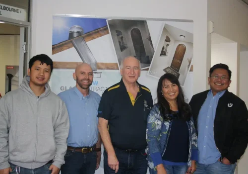 A group of five people, known for their exceptional service, stand indoors in front of a display board featuring images of doors and windows. They are smiling and dressed casually.