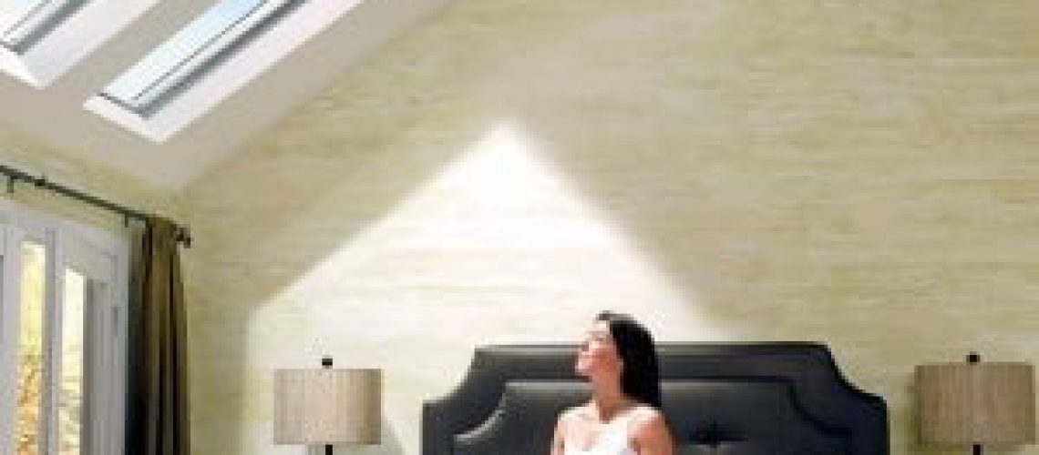 A woman sits on a bed in a bright, modern bedroom with skylights that showcase the benefits of skylights for your bedroom, and two bedside lamps.