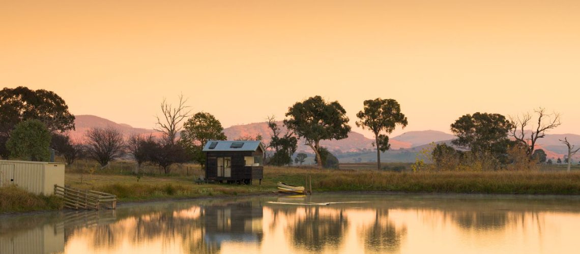 A serene lakeside scene at sunset with a small cabin featuring skylights in tiny homes, surrounded by trees and hills in the background.