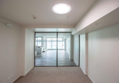 A room with beige walls and carpet leads to another room with wooden flooring, separated by large glass doors. A round ceiling light, complemented by a Solatube skylight, illuminates the area.