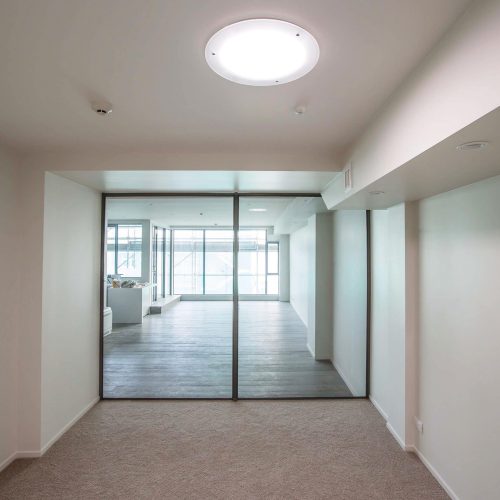 A room with beige walls and carpet leads to another room with wooden flooring, separated by large glass doors. A round ceiling light, complemented by a Solatube skylight, illuminates the area.