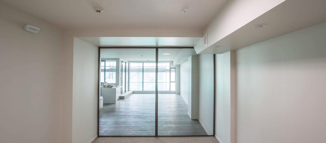 A room with beige walls and carpet leads to another room with wooden flooring, separated by large glass doors. A round ceiling light, complemented by a Solatube skylight, illuminates the area.